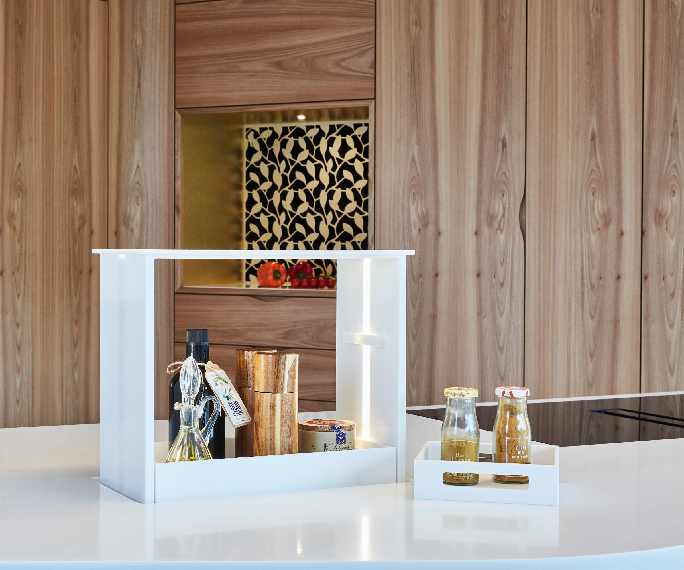 A sleek white pop-up kitchen storage unit rises from the worktop, holding oils, spices and cooking essentials. Wood-grain cabinets and patterned splashback tiles frame the scene, blending clever concealed functionality with elegant modern design.