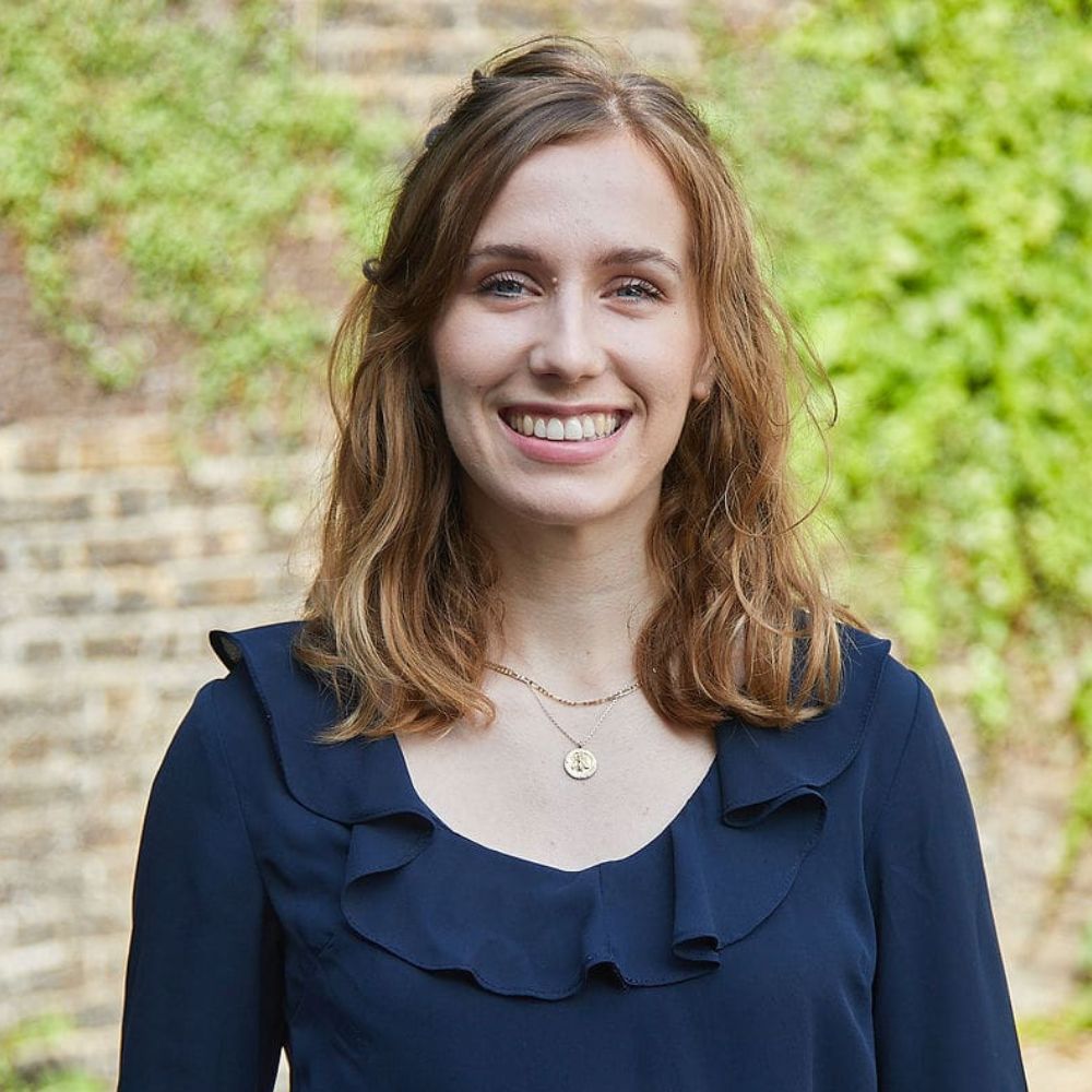 A woman with shoulder-length light brown hair smiling at the camera. She is wearing a navy blouse with a ruffled neckline and a gold necklace, standing outdoors in front of a stone wall and greenery.
