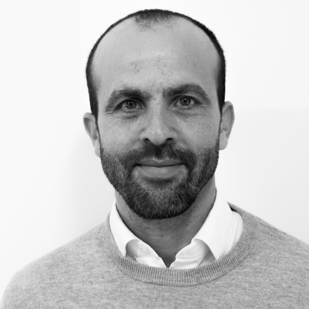 A man with a closely shaved head and trimmed beard wearing a light grey jumper over a white collared shirt, smiling softly against a plain white background.