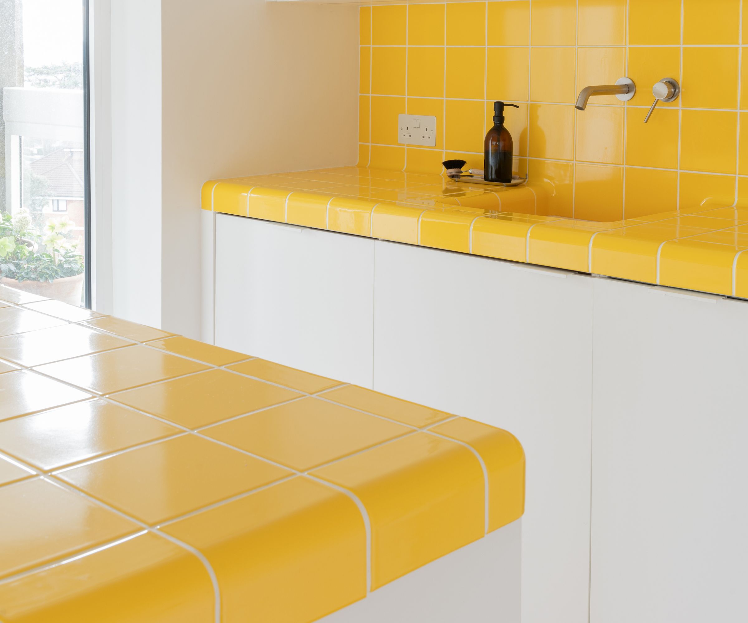 Yellow tiled kitchen worktop and splashback with integrated sink and white base units in a bright, modern kitchen.