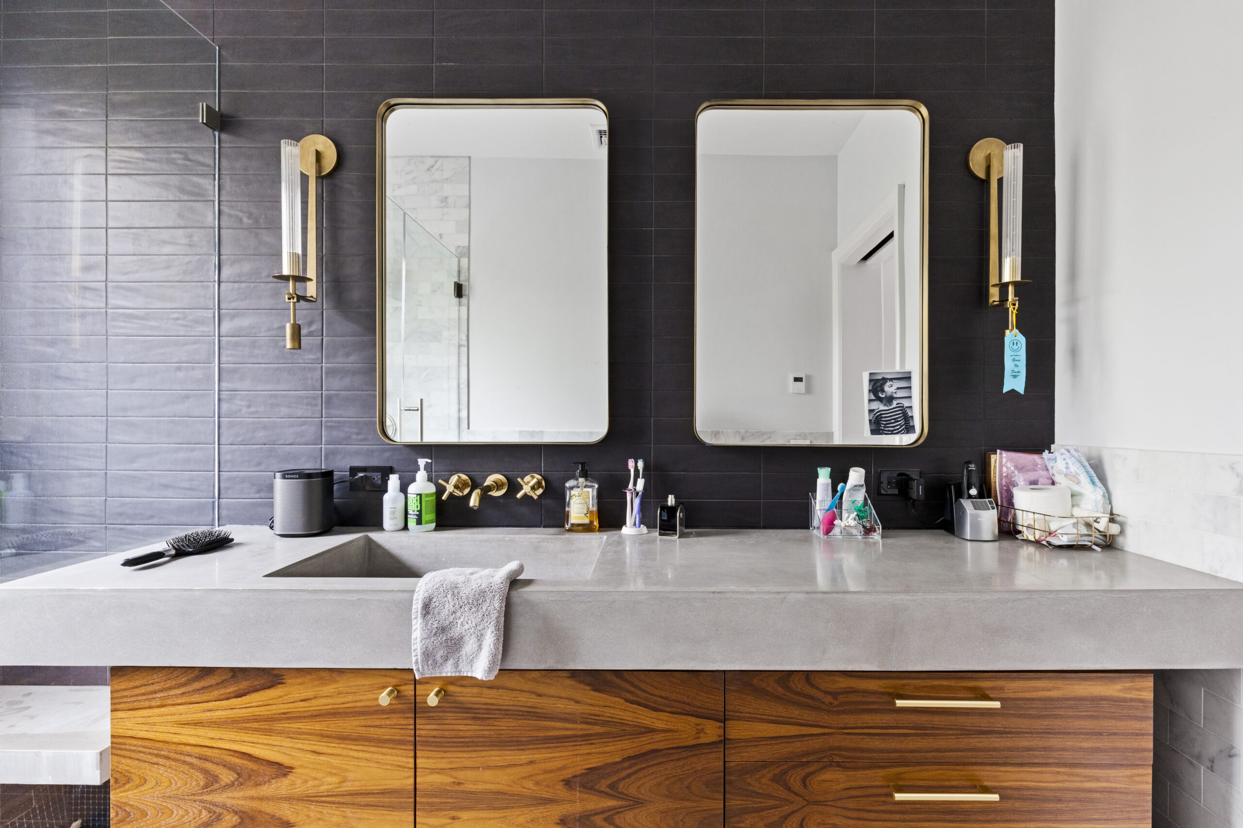 An updated bath showcasing a walnut bathroom vanity, concrete counter, brass pulls, and twin mirrors on charcoal tile.