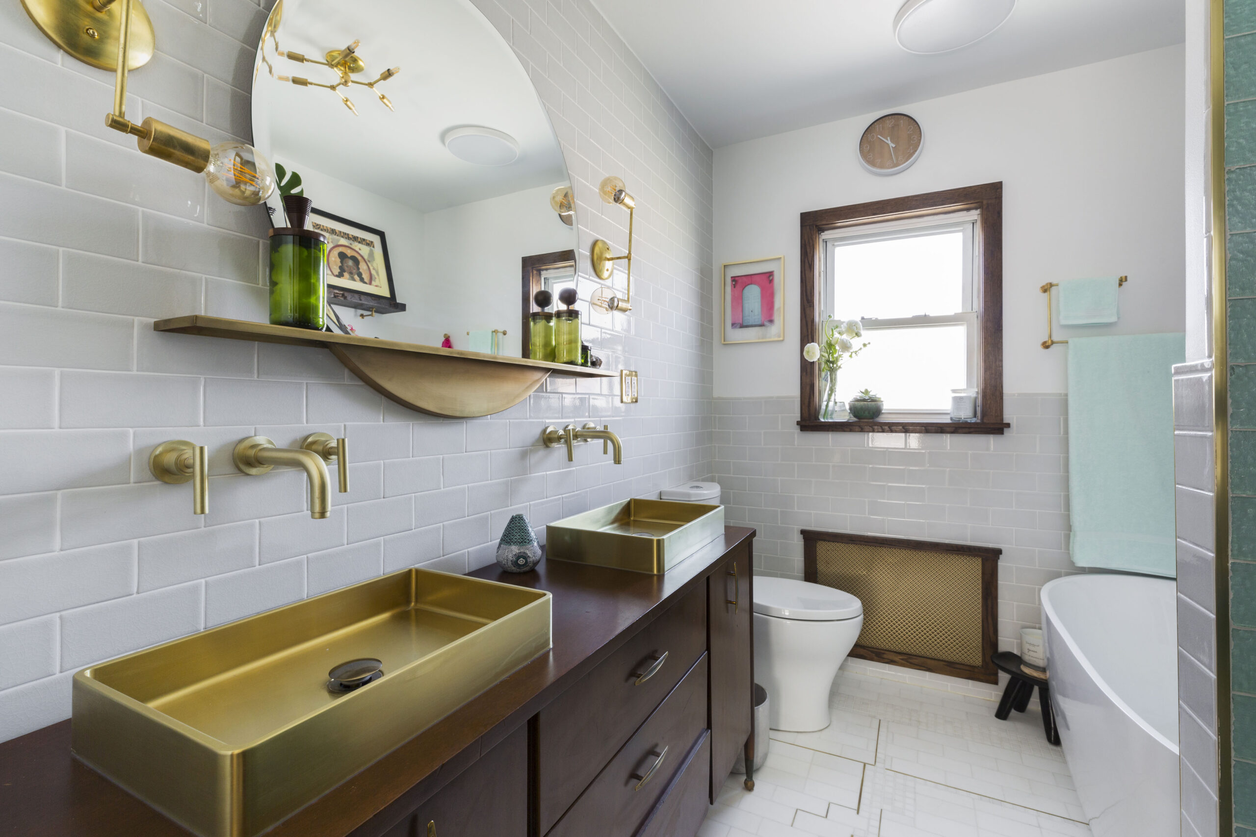 A remodeled bath showing a dark wood bathroom vanity, twin brass trough sinks, wall faucets, and an arched mirror.