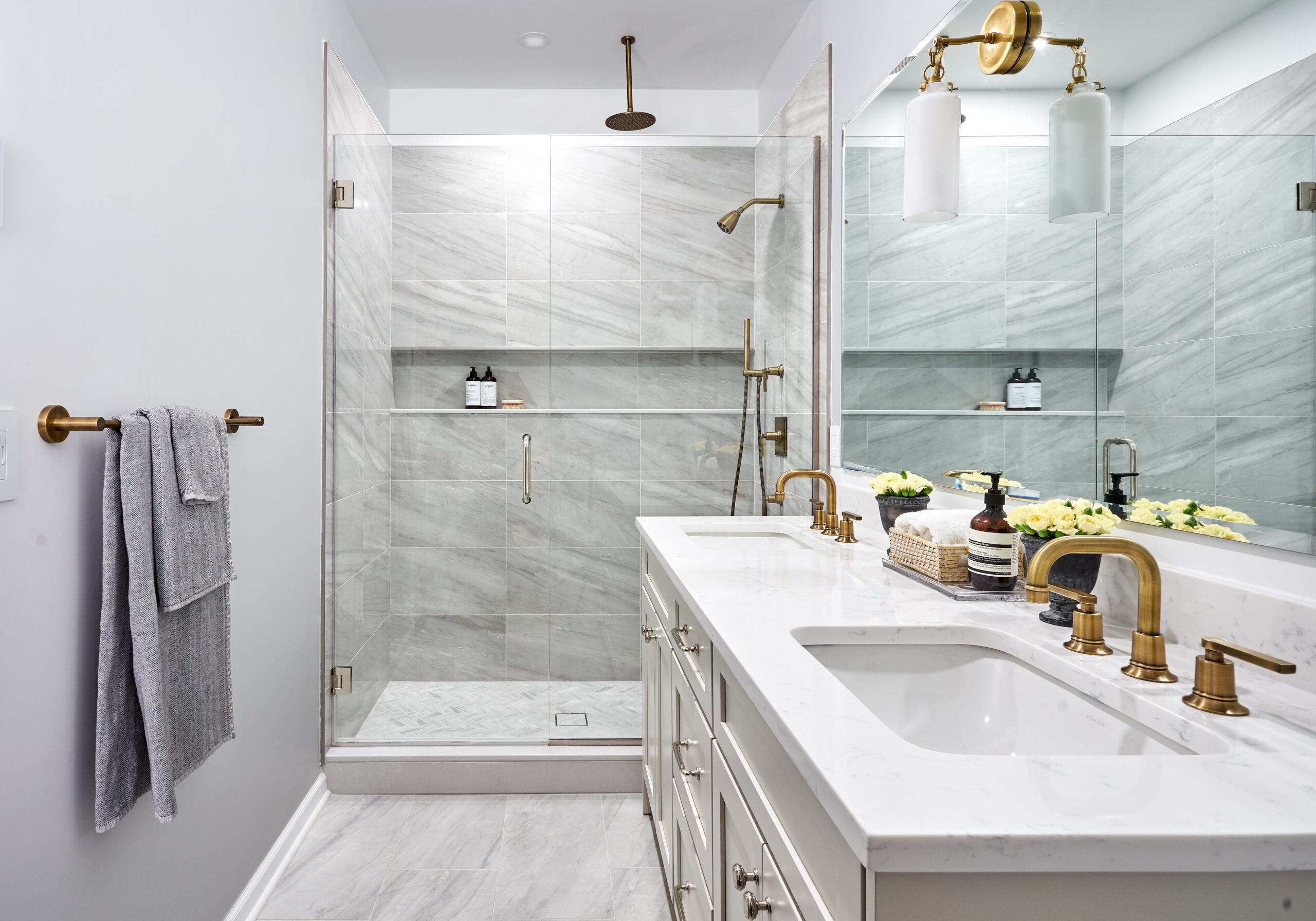 A renovated bath features a white bathroom vanity, quartz top, brass fixtures, and a glass shower in marble-look tile.