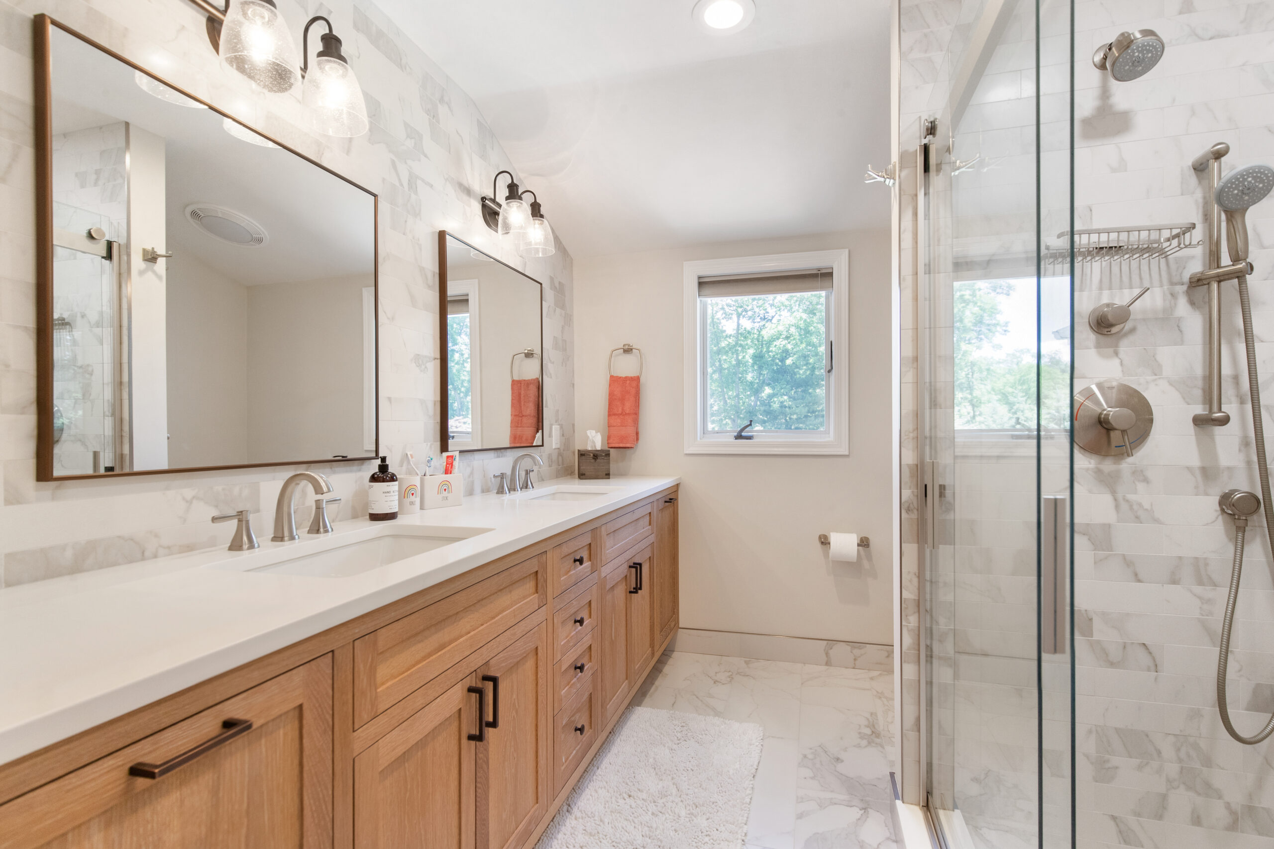An updated bath with a light oak bathroom vanity, double sinks, mirrors, and a glass shower with marble tile.