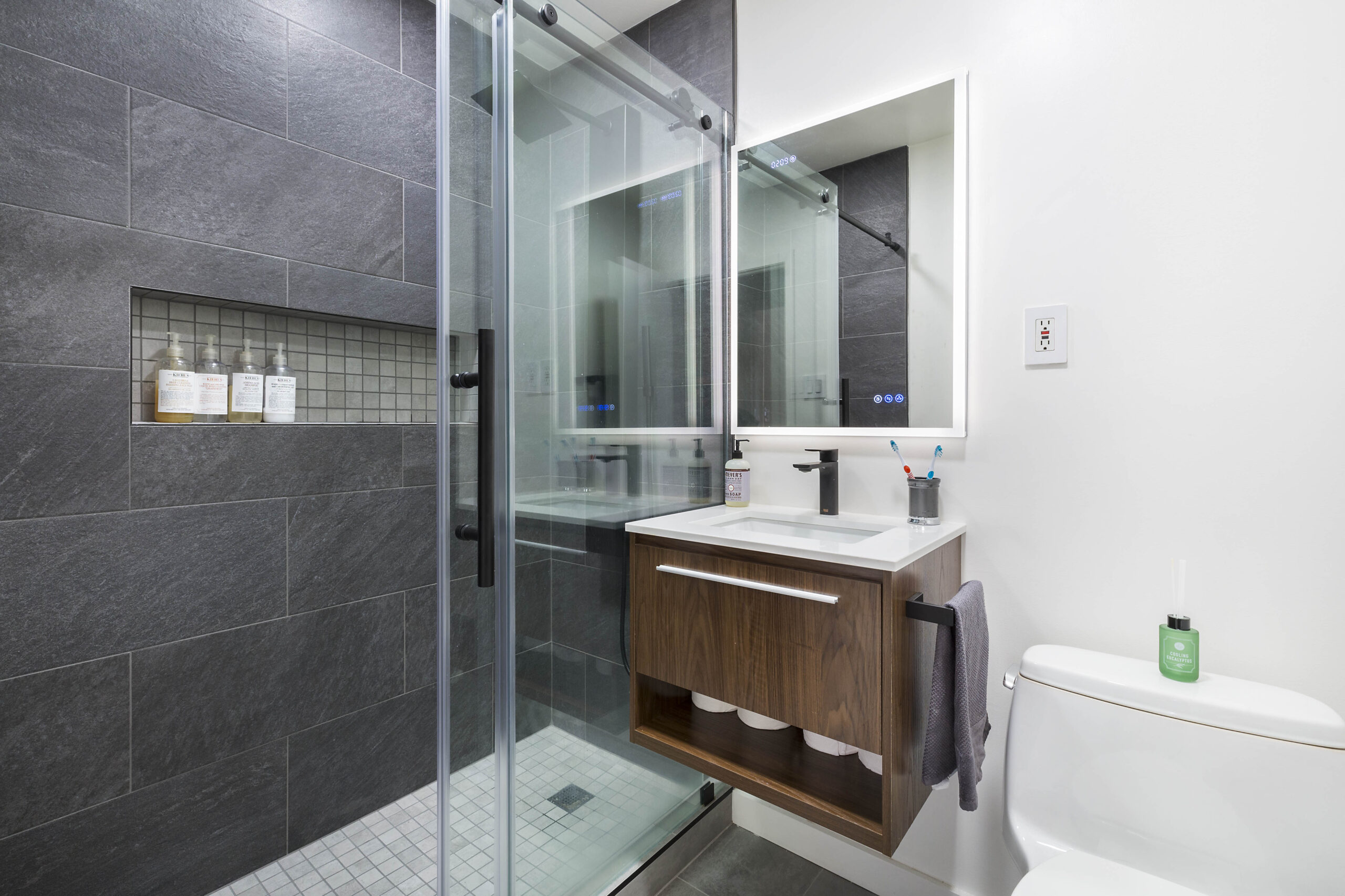 A refreshed bath featuring a floating bathroom vanity, backlit mirror, black faucet, and glass shower with dark tile.