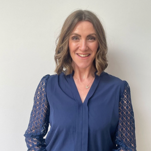 female with shoulder length hair wearing blue blouse stood against wall