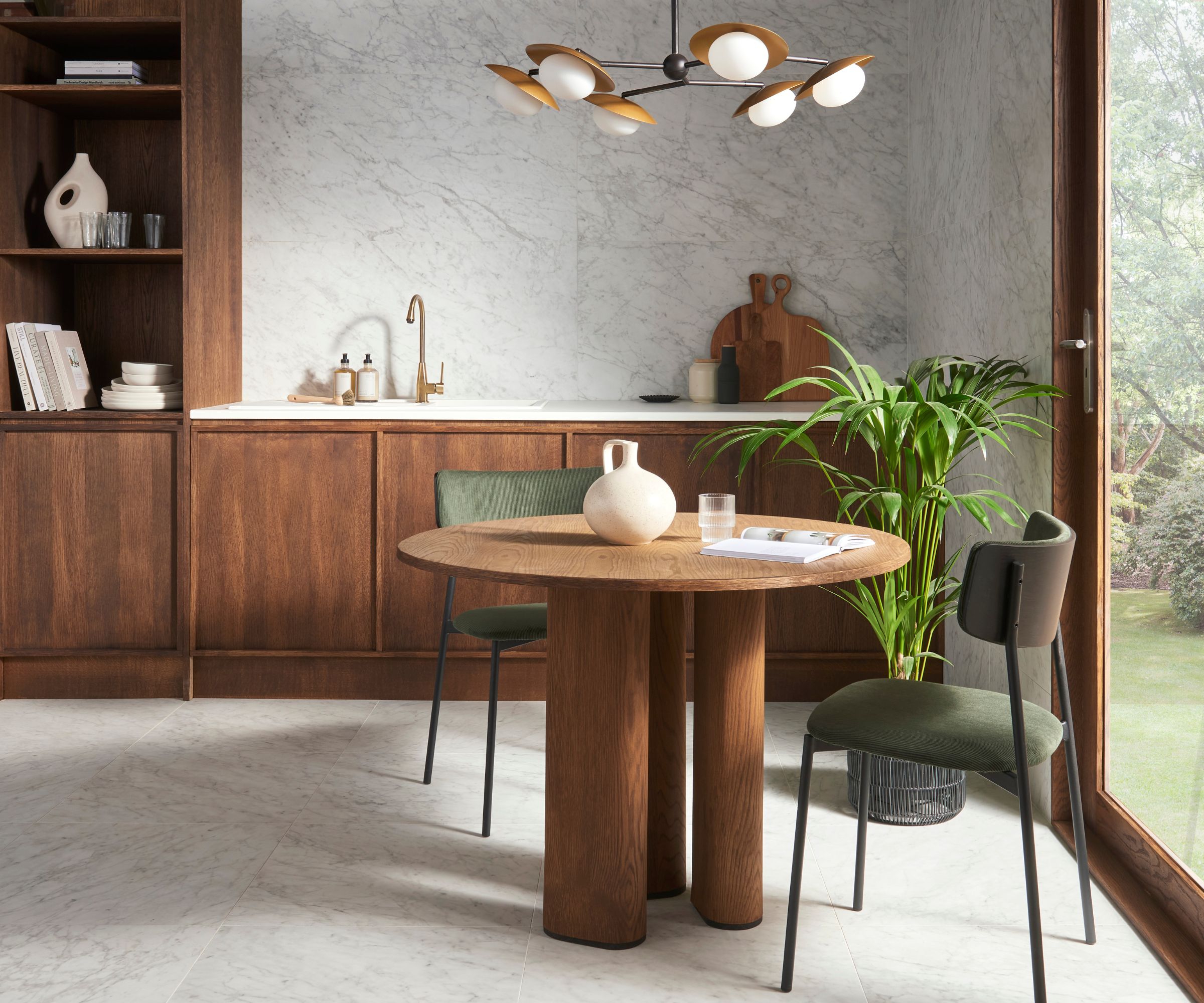 Compact kitchen-dining space with dark wood units, marble backsplash, round wooden table, green chairs and a large plant by a glass door.
