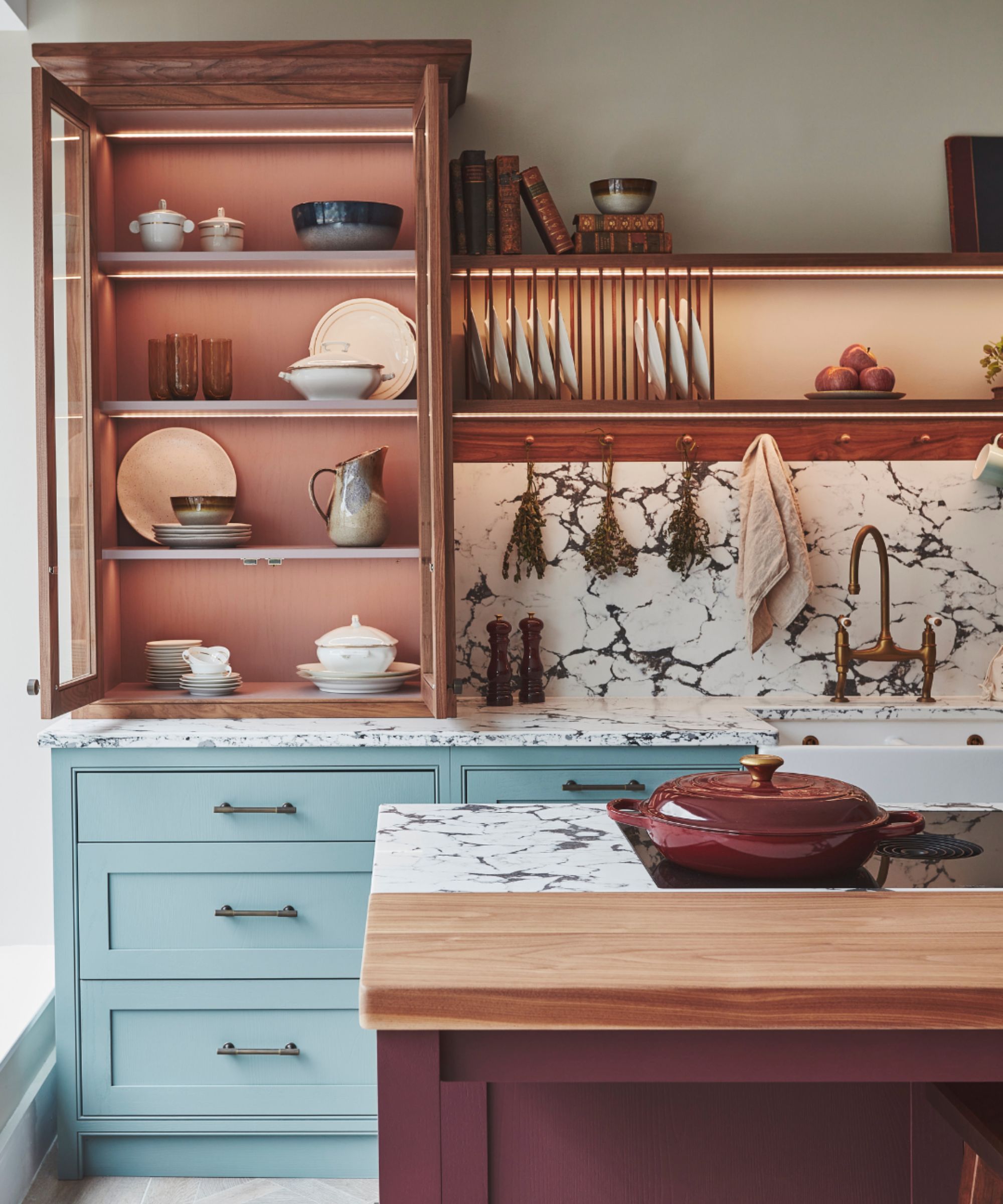 Pale blue kitchen with marble worktops, open wooden shelving lit by warm LEDs, a plate rack, brass tap and a wooden-topped island in front.