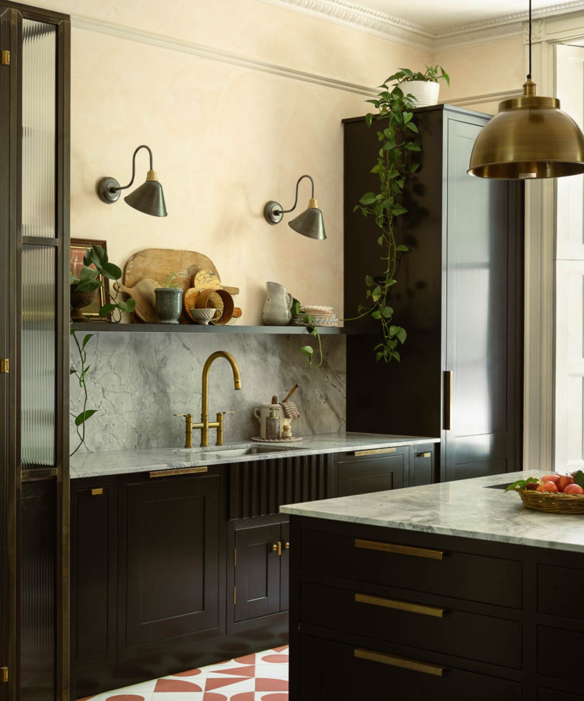 A dark, moody kitchen with deep brown cabinetry and brass handles, featuring a marble splashback and worktops. Open shelves display ceramic bowls, cups and wooden boards, with trailing green plants placed along the shelf and on top of tall units.