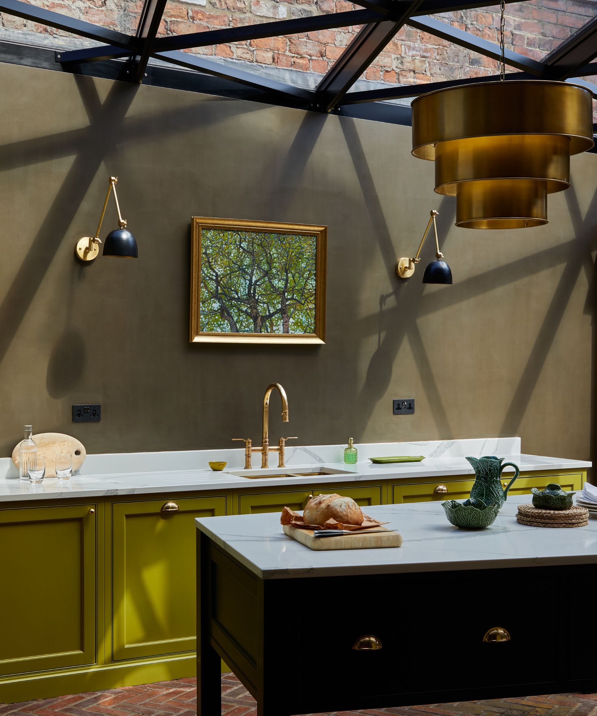 A characterful kitchen with olive-green cabinetry, brass details and a glass roof, blending natural light with rich, heritage-inspired colour.